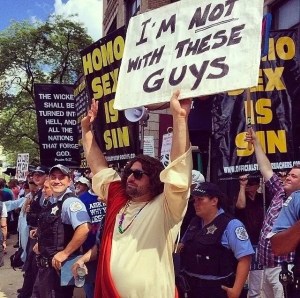 Man dressed as Jesus stands in front of a group of anti-gay protestors. Jesus-guy holds a sign that says, “I'm not with these guys.”